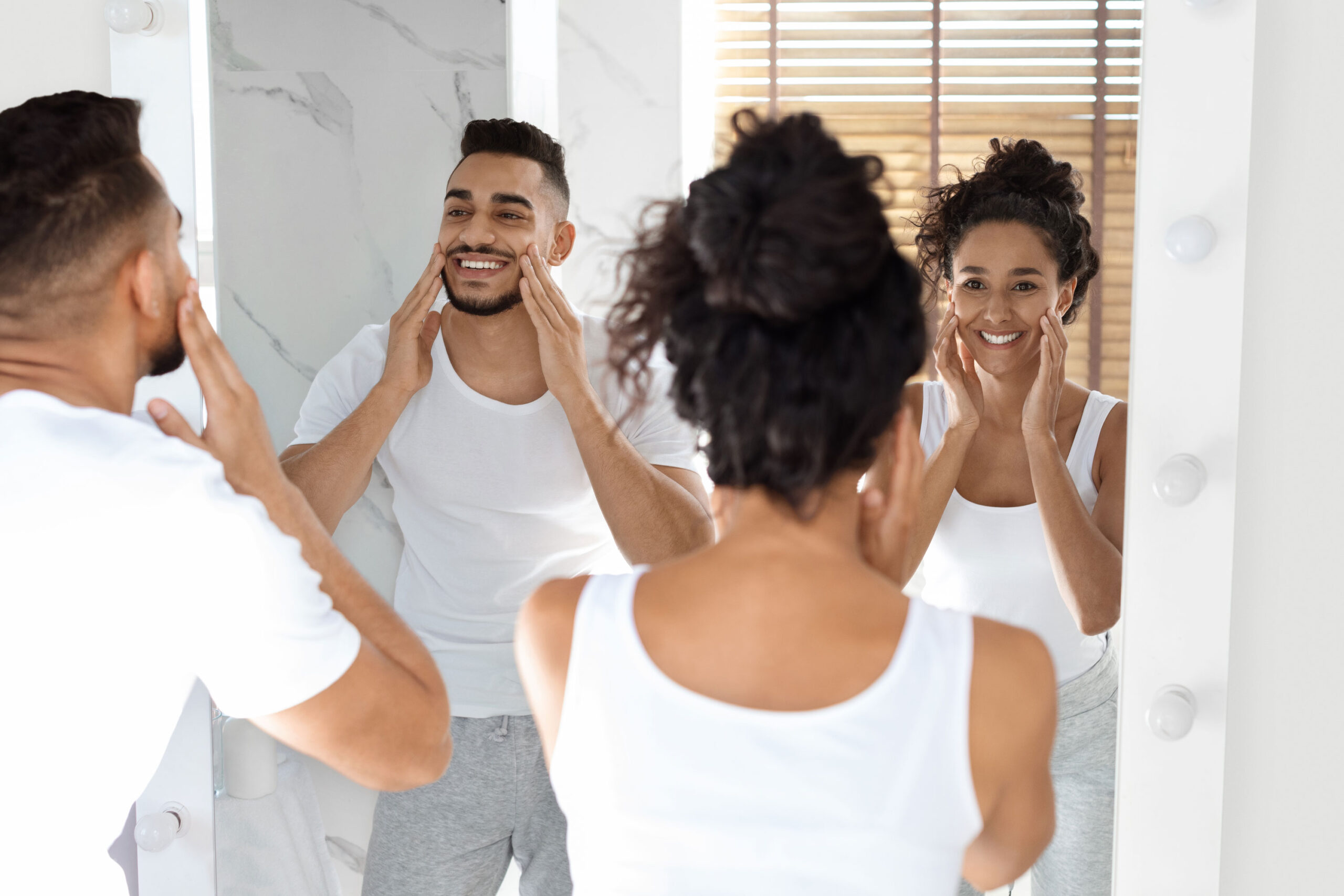 A man and a woman, both wearing white tops, stand in front of a bathroom mirror smiling and touching their faces.